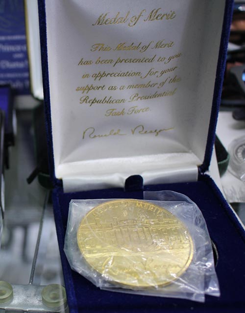 A gold coin in a plastic and blue box, which has a label "Medal of Merit"