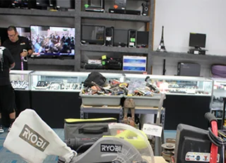 A pawn shop with accessories on display, some electronics displayed behind the counter, 1 staff and a customer, some tools in a table