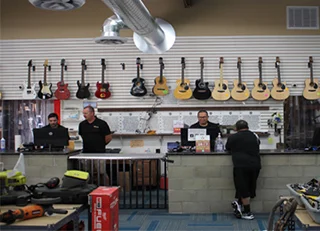 A counter of a pawnshop with 3 staffs and 1 customer, guitars hanging on the wall, some tools in the table